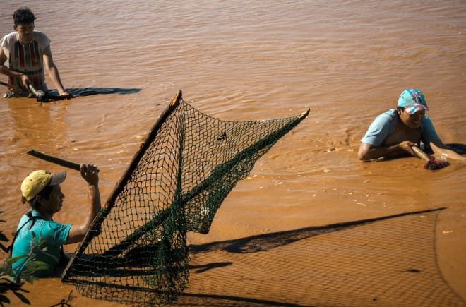 Três homens pescando, em pé, com água até a cintura, em um rio marrom