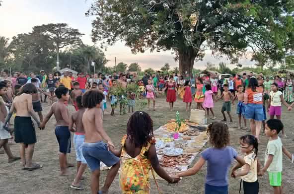 Dezenas de pessoas, entre mulheres, homens, crianças e idosos, estão em círculo em torno de uma toalha estendida no chão com alimentos, sementes e outros produtos da terra