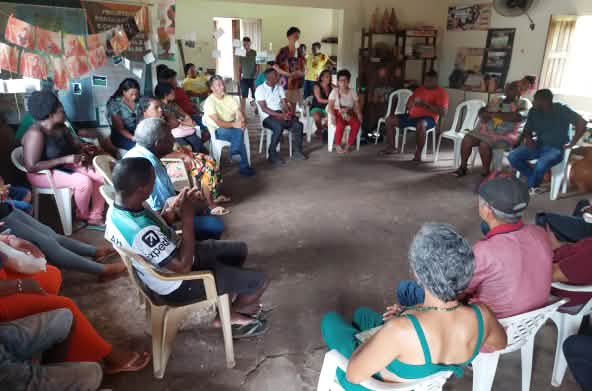 Cerca de 25 pessoas sentadas em roda estão conversando sobre impactos de projeto porto-ferroviário. Estão em uma sala com algumas prateleiras de livros e fotos nas paredes.