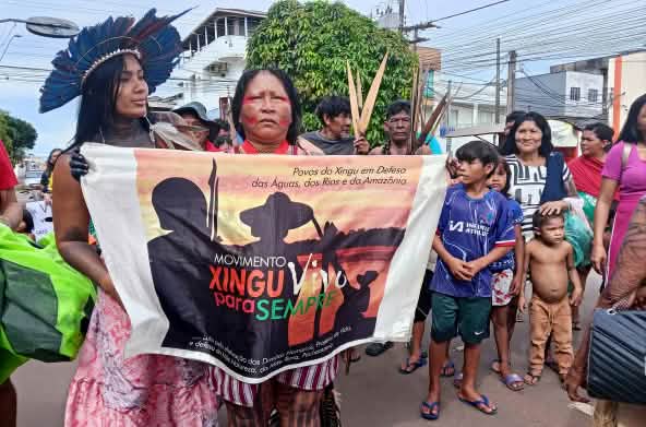 Duas mulheres indígenas seguram uma faixa de protesto. Atrás delas há homens adultos e crianças