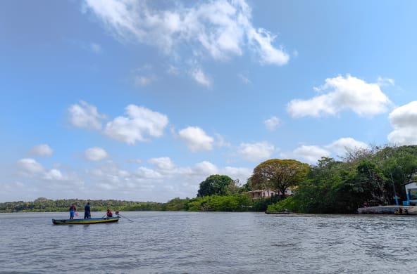 Sob o céu azul, um pequeno barco navega pelo mar com três pescadores. À direita estão pequenas casas no meio de uma floresta.