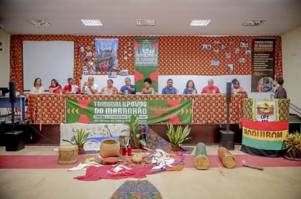 Doze pessoas estão sentadas atrás de uma mesa enfeitada com pano colorido e cartaz escrito Tribunal dos Povos do Maranhão