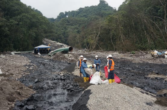 Grupo de petroleiros trabalhando entre dois fluxos de petróleo pesado, que vão se derramando morro abaixo em direção à floresta tropical, na encosta coberta de cascalho. No fundo, vê-se uma parte da tubulação do oleoduto.