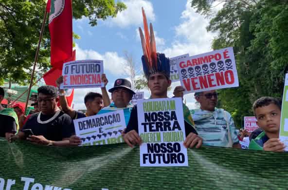 Indígenas marcham em uma fileira e elevam pôsteres com textos como: "Garimpo é veneno" e "O futuro é indígena"