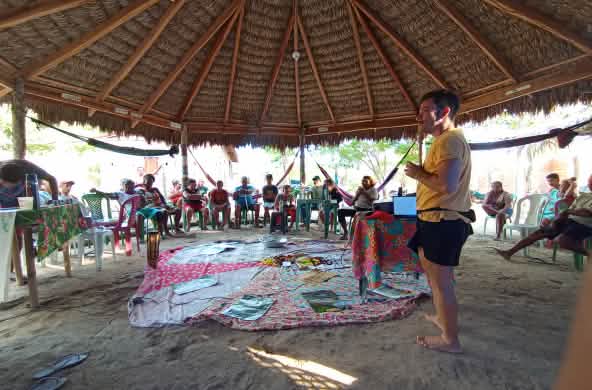 Sentados em círculo em cadeiras, sob uma tenda de madeira e palha, homens e mulheres conversam com um homem que está em pé, ouvindo-os