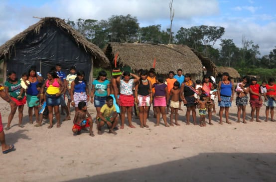 Pessoas da etnia Ka’apor em fila dupla diante de duas cabanas na floresta