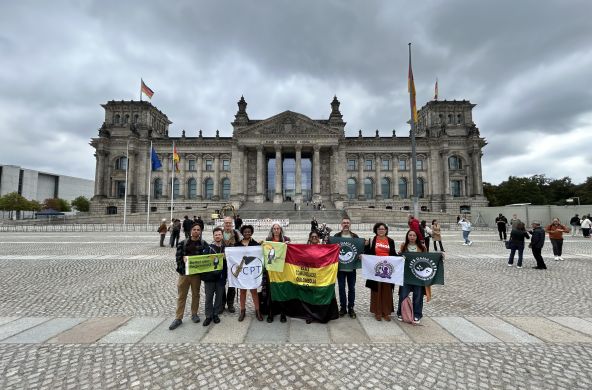 Nove pessoas em pé, segurando bandeiras com logos e nomes de organizações, posam para foto em frente à sede do parlamento alemão