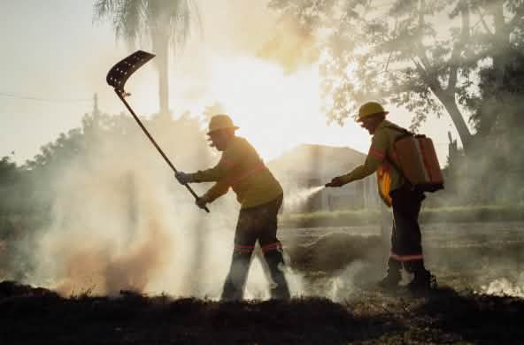 Em meio à fumaça, um homem combate o incêndio com um extintor de incêndio, enquanto outro carrega uma mochila com água e uma mangueira manual