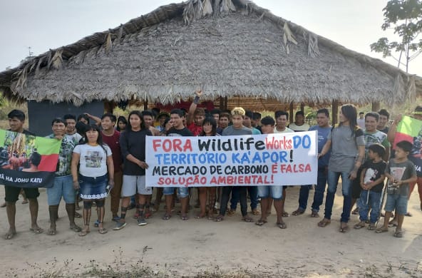 Mais de 30 pessoas do povo Ka'apor estão em pé, diante de uma casa com pilares de madeira e telhado de palha. As pessoas seguram uma faixa de protesto com a frase: "Fora Wildlife Works do território Ka'apor! Mercado de carbono é falsa solução ambiental!"