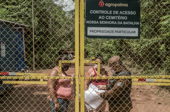 Duas mulheres sendo controladas por um guarda por trás de um portão gradeado - Texto inscrito na placa da empresa: Agropalma - Controle de Acesso ao Cemitério Nossa Senhora da Batalha. PROPRIEDADE PARTICULAR