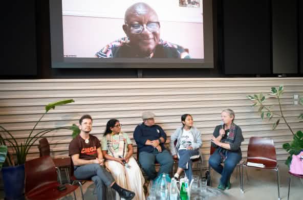 Cinco pessoas sentadas conversam. Acima delas, em um telão, um homem participa da conversa virtualmente