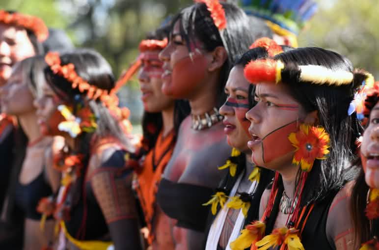 Protesto indígena no Acampamento Terra Livre em Brasília 2024 Oito mulheres indígenas, lado a lado, com enfeites de plumas e penas em suas cabeças, brincos de penas amarelas e vermelhas, e pintura vermelha e preta no rosto