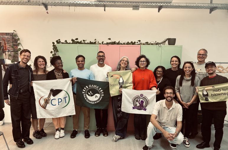 Onze pessoas em pé e uma agachada sorriem para a foto segurando bandeiras com nomes e logos de organizações