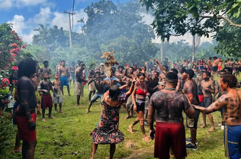 Roda com dezenas de indígenas com seus corpos pintados de preto e um galo voando em meio às pessoas