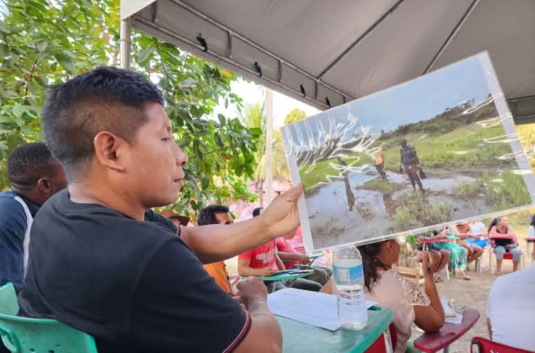 Homem Awa Guajá sentado observa uma foto. Ele está em uma cadeira em meio a um círculo com outras pessoas também sentadas