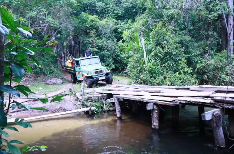Caminhão carregando pessoas na carroceria se prepara para cruzar ponte de tábuas de madeira sobre rio