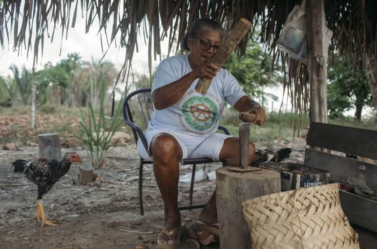 Mulher de camiseta e bermuda brancas está sentada quebrando, sobre um tronco de madeira, um coco babaçu. Ela usa uma cunha e uma manceta