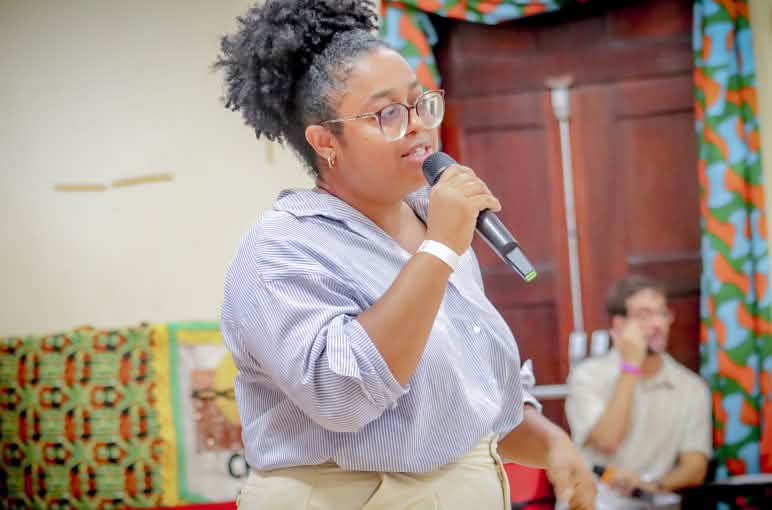 Mulher negra com cabelos presos, óculos de grau, camisa azul claro e calça bege segura microfone com a mão direita diante da boca enquanto fala