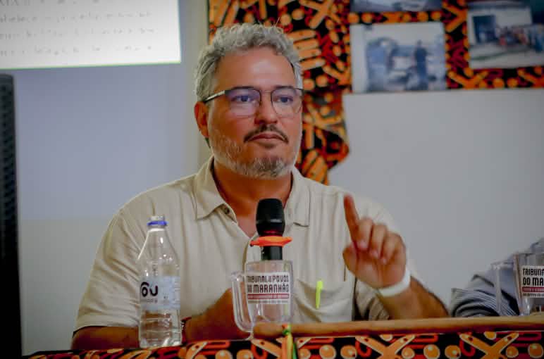 Homem branco, barba e cabelos grisalhos, usando óculos de grau e camisa bege está sentado e segura microfone em uma das mãos