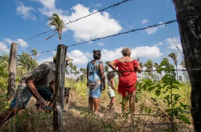 Um homem passa por baixo de uma cerca de arame farpado enquanto duas mulheres e uma criança, já depois da cerca, observam babaçual