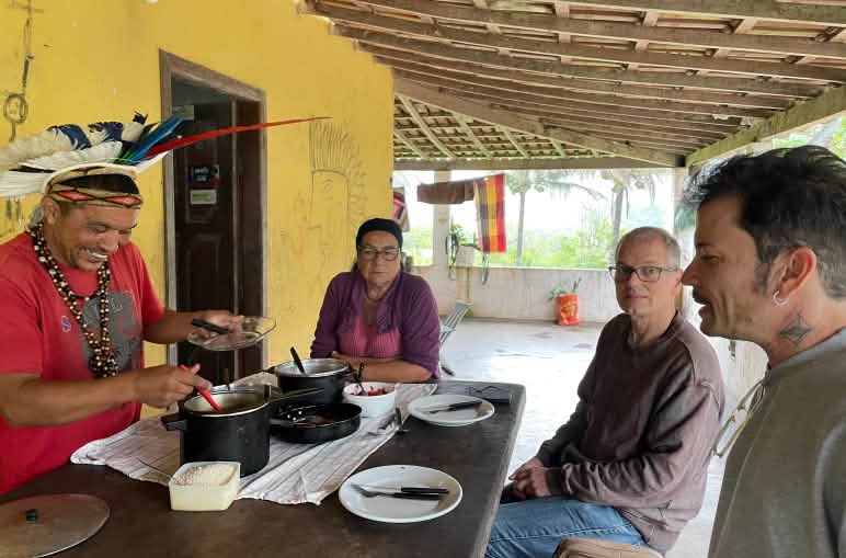 Mãdy Pataxó, adornado com um cocar de penas, sua mãe, Felipe Sabrina e Klaus Schenck, ativistas de Salve a Floresta sentados em torno de uma mesa de madeira preparada para o almoço