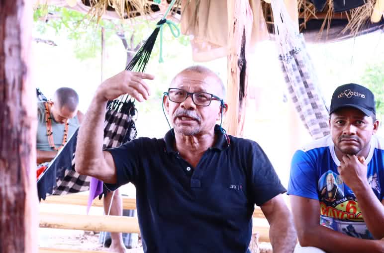 Homem usando óculos de grau e camisa preta está sentado, falando, gesticulando o braço direito
