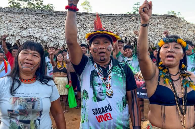 Três indígenas Ka'apor - duas mulheres e um homem - com os punhos erguidos em frente a um abrigo coberto com folhas de palmeira