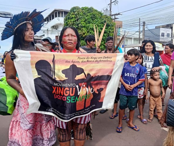 Duas mulheres indígenas seguram uma faixa de protesto. Atrás delas há homens adultos e crianças