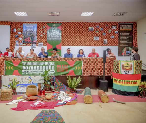 Doze pessoas estão sentadas atrás de uma mesa enfeitada com pano colorido e cartaz escrito Tribunal dos Povos do Maranhão