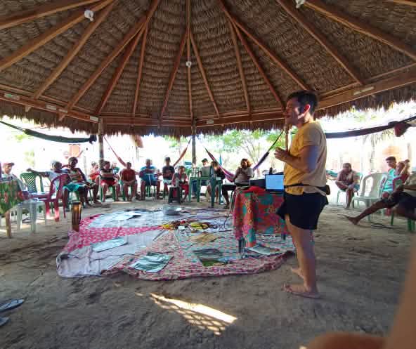 Sentados em círculo em cadeiras, sob uma tenda de madeira e palha, homens e mulheres conversam com um homem que está em pé, ouvindo-os
