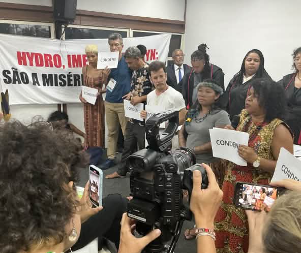 Mais de 10 pessoas estão em pé diante de uma platéia. Algumas pessoas seguram uma placa onde está escrito "condeno", em referência ao veredito do júri