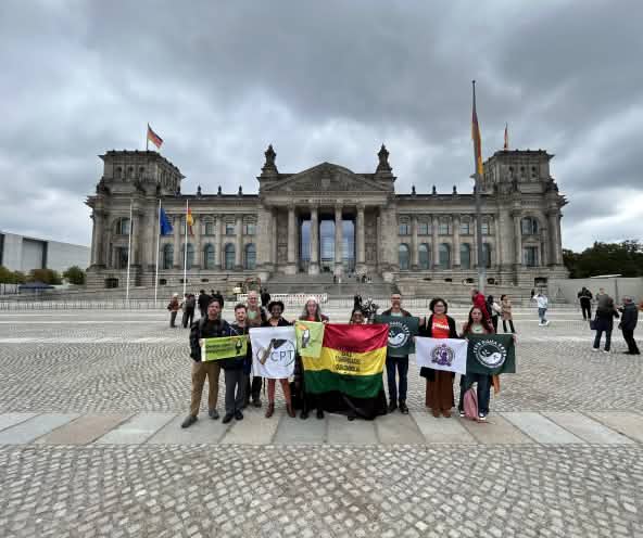 Nove pessoas em pé, segurando bandeiras com logos e nomes de organizações, posam para foto em frente à sede do parlamento alemão