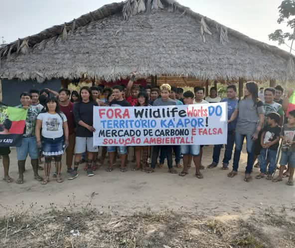 Mais de 30 pessoas do povo Ka'apor estão em pé, diante de uma casa com pilares de madeira e telhado de palha. As pessoas seguram uma faixa de protesto com a frase: "Fora Wildlife Works do território Ka'apor! Mercado de carbono é falsa solução ambiental!"