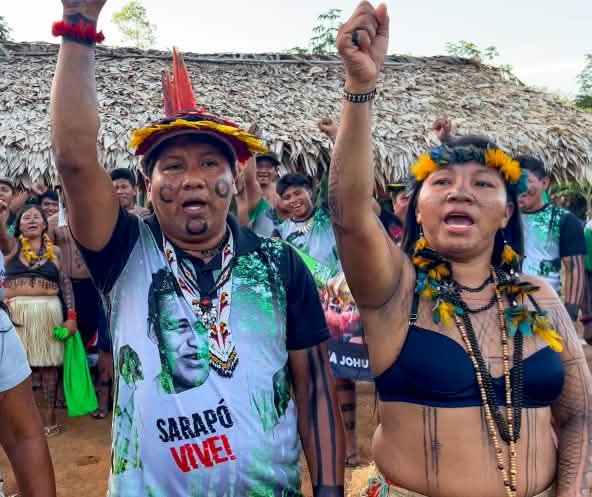 Três indígenas Ka'apor - duas mulheres e um homem - com os punhos erguidos em frente a um abrigo coberto com folhas de palmeira