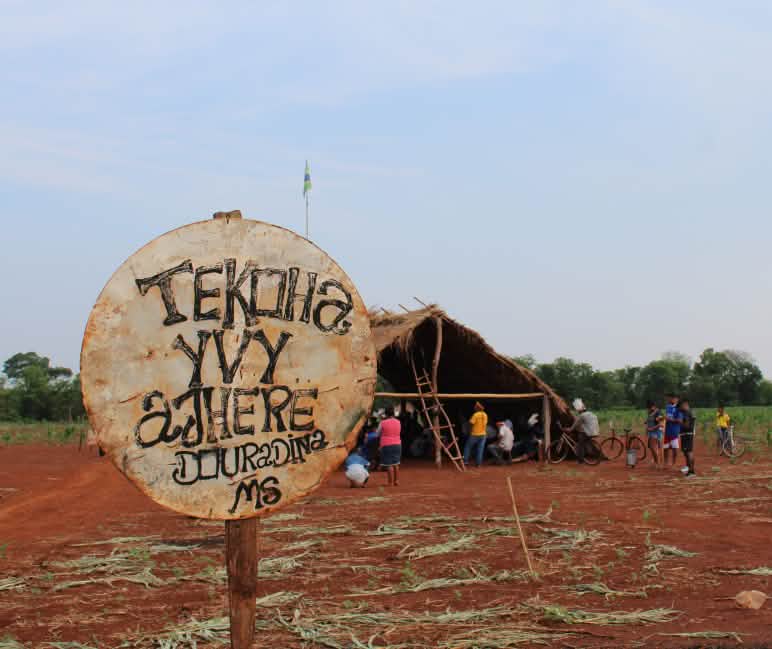 Um sinal pintado à mão em primeiro plano e, em segundo plano, um abrigo improvisado com pessoas num antigo terreno agrícola