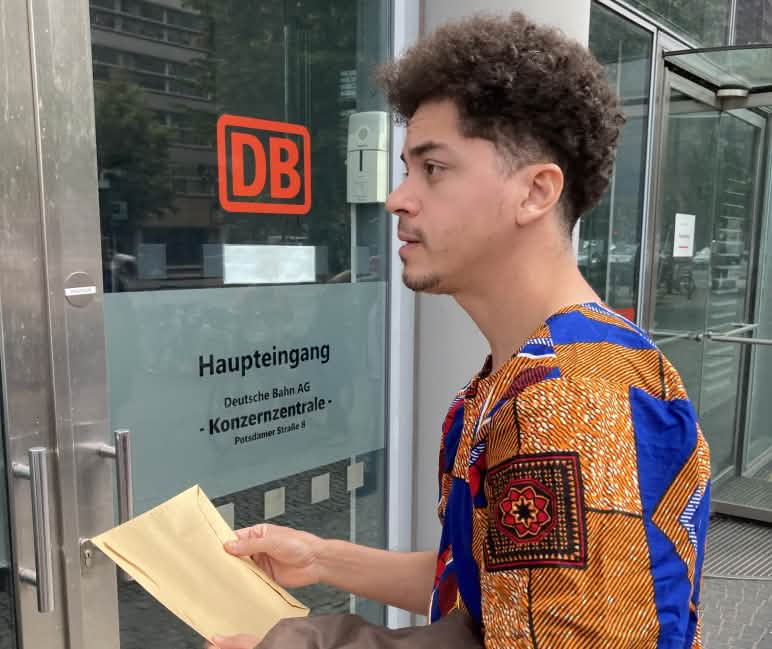 Um jovem está parado com um envelope em frente a uma porta de vidro com a etiqueta DB Entrada principal da sede do Grupo