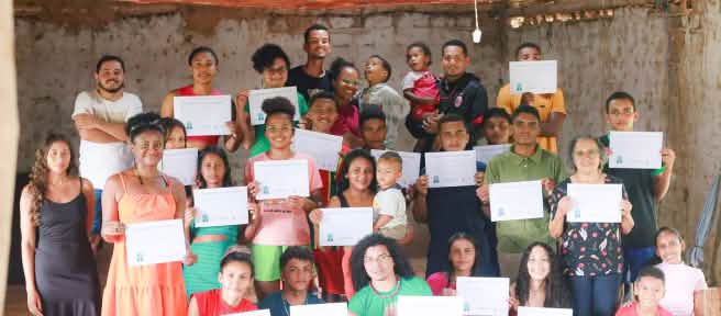 31 pessoas, entre mulheres, homens, jovens e crianças estão em pé, algumas agachadas, em uma sala com paredes brancas ao fundo. A maioria das pessoas segura diante do peito, com as duas mãos, um diploma de conclusão da oficina escrito em folha branca.