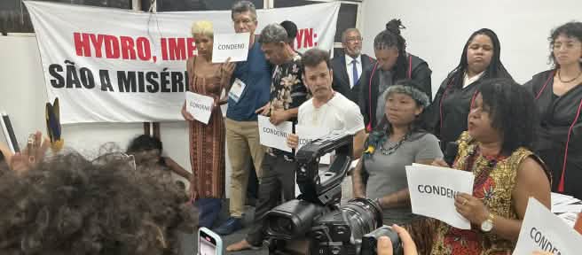 Mais de 10 pessoas estão em pé diante de uma platéia. Algumas pessoas seguram uma placa onde está escrito "condeno", em referência ao veredito do júri