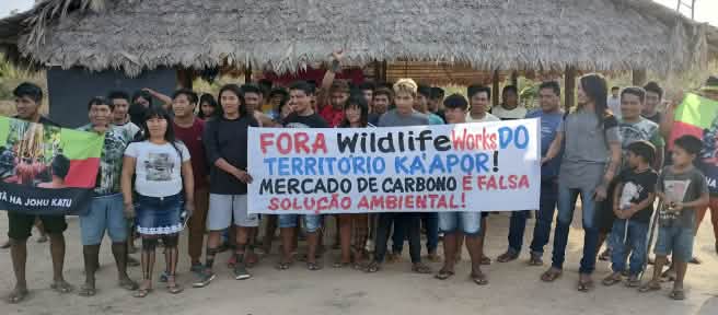 Mais de 30 pessoas do povo Ka'apor estão em pé, diante de uma casa com pilares de madeira e telhado de palha. As pessoas seguram uma faixa de protesto com a frase: "Fora Wildlife Works do território Ka'apor! Mercado de carbono é falsa solução ambiental!"