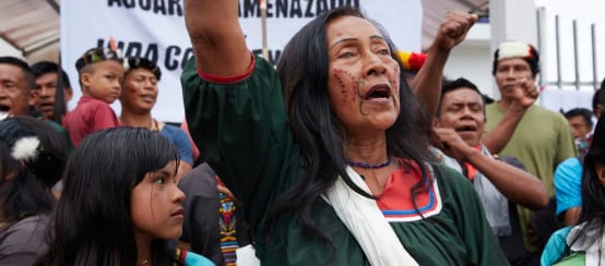 Moradores nativos, mulheres, crianças e homens diante de uma faixa, fazendo uma manifestação