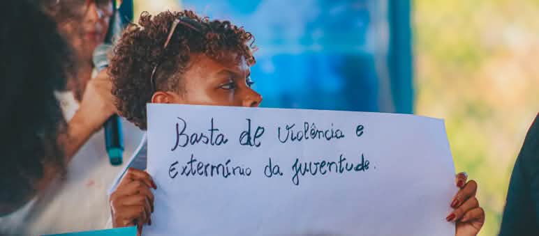 Mulher jovem segura cartaz onde está escrito "Basta de violência e extermínio da juventude"