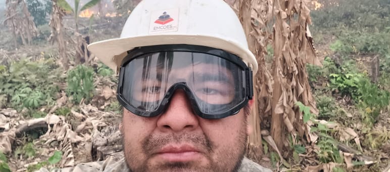 Homem de capacete e óculos de proteção olhando para a câmara, enquanto ao fundo, há arbustos queimando e fumaça subindo