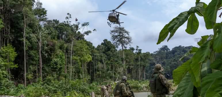 Dois agentes do IBAMA, armados e vestidos de uniformes de camuflagem, sob um helicóptero sobrevoando área desmatada na floresta