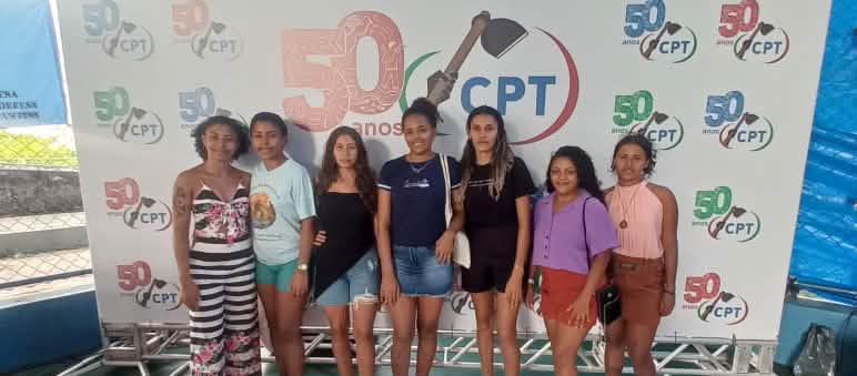 Sete mulheres jovens aparecem em pé, perfiladas e sorrindo para a foto. Atrás delas há um banner com a logomarca escrito 50 anos CPT