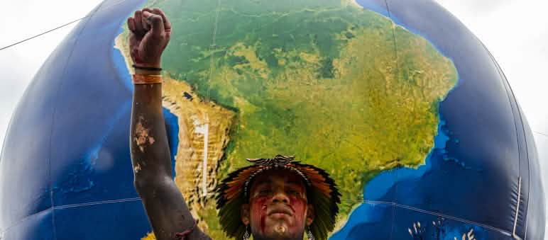 Indígena brasileiro com o punho erguido em frente a um globo mostrando a América do Sul