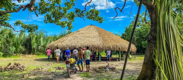 Ao fundo, casa redonda construída na aldeia Tabarelzinho após a retomada. Espaço é usado para reuniões e outras atividades coletivas Tenda redonda coberta com palhas está no meio da floresta de palmeiras. Algumas pessoas estão embaixo da tenda, outras estão chegando no local. Uma moto está estacionada na frente da tenda.