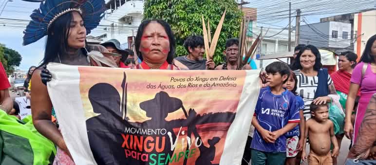 Duas mulheres indígenas seguram uma faixa de protesto. Atrás delas há homens adultos e crianças