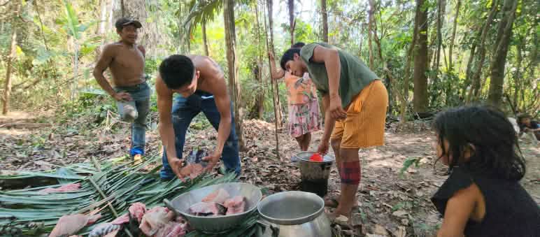 Cinco pessoas estão no meio da floresta preparando alimentos em folhas e panelas colocadas no solo