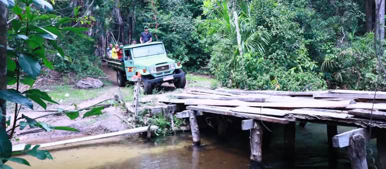 Caminhão carregando pessoas na carroceria se prepara para cruzar ponte de tábuas de madeira sobre rio