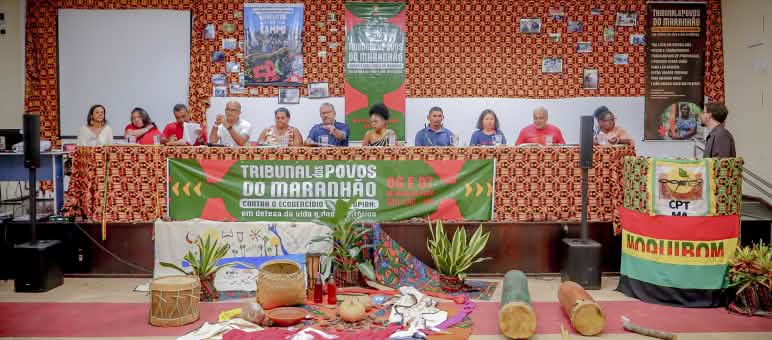 Doze pessoas estão sentadas atrás de uma mesa enfeitada com pano colorido e cartaz escrito Tribunal dos Povos do Maranhão